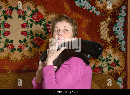 Close-up halblängen Porträt ältere Dame in hell Magenta Bluse, die auf ihrer Schulter die schwarze Katze auf handgemachte print Teppich staatlich hält Stockfoto