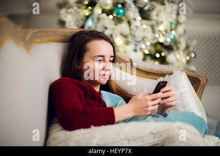 Junge Frau beim Auschecken ihre SMS-Nachrichten auf dem Handy als She Liyng Sofa vor dem Weihnachtsbaum zu Hause. Stockfoto