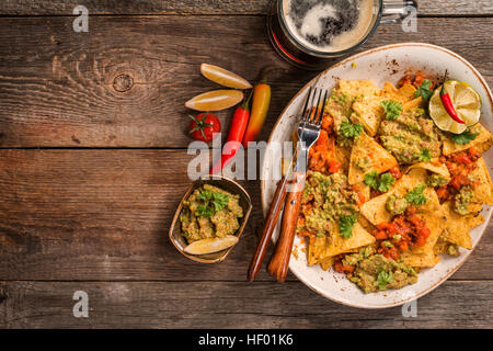 Gelber Mais Nacho, die Chips garniert mit Hackfleisch, Guacamole, Käse überbacken, Paprika und Petersilie Blätter in Platte mit Bier auf Holztisch, Ansicht von oben Stockfoto