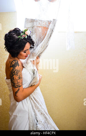 Schöne Brünette lächelnde Braut posiert mit seiner Hochzeit Spitzenkleid in der hand und Spiegel zu betrachten. Emotionale Mädchen tragen weiße Blumen im Haar. Stockfoto