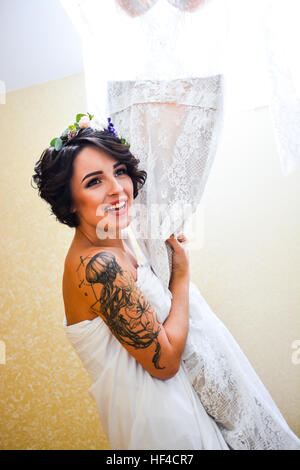 Schöne Brünette lächelnde Braut posiert mit seiner Hochzeit Spitzenkleid in der hand und Spiegel zu betrachten. Emotionale Mädchen tragen weiße Blumen im Haar. Stockfoto