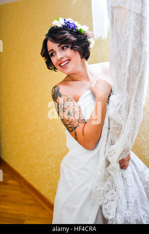 Schöne Brünette lächelnde Braut posiert mit seiner Hochzeit Spitzenkleid in der hand und Spiegel zu betrachten. Emotionale Mädchen tragen weiße Blumen im Haar. Stockfoto