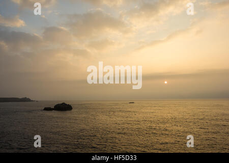 Die Kugel der Sonne steigt durch niedrige Wolkendecke über ein ruhiges Meer und Felsen Stockfoto