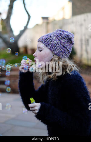 Junge, blondes Mädchen schwarz gekleidet mit einem lila Hut auf Schläge Bläschen an einem kalten Wintertag. Stockfoto