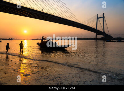 Silhouette Straßenkinder spielen bei Sonnenuntergang am Ufer des Fluss Hooghly mit Vidyasagar Setu (Brücke) auf dem Hintergrund. Stockfoto