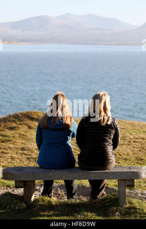 Zwei weibliche Freunde sitzen Seite an Seite mit dem Rücken auf eine Bank stellte sich heraus, dass mit Blick auf das Meer und den Snowdonia National Park von llanddwyn Island am Strand Vinderup, Anglesey Stockfoto