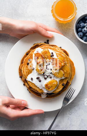 Pfannkuchen zum Frühstück. Pfannkuchen mit karamellisierten Bananen, Joghurt und Schokolade. Frau Hände Halteplatte mit Stapel von Pfannkuchen Stockfoto