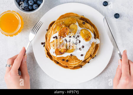 Pfannkuchen mit karamellisierten Bananen, Joghurt und Schokolade-Chips auf weißen Teller. Mädchen essen Pfannkuchen zum Frühstück Stockfoto