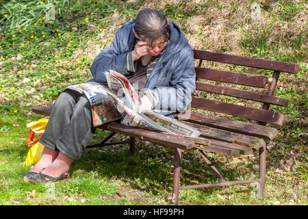 Eine ältere Frau liest Zeitungen in einem Stadtpark auf einer Bank, ohne Sehvermögen Stockfoto