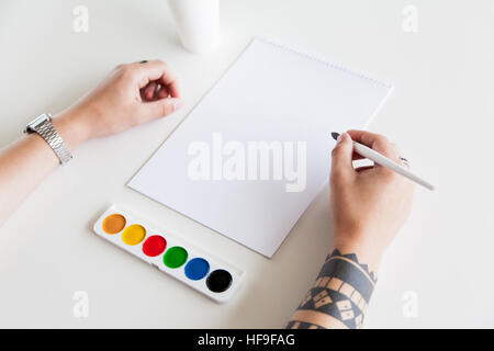 Die Hand des Mannes mit einer Tätowierung halten Sie den Pinsel vor leerem Papier und Farben auf weißem Hintergrund. Mock-up-Branding. Stockfoto