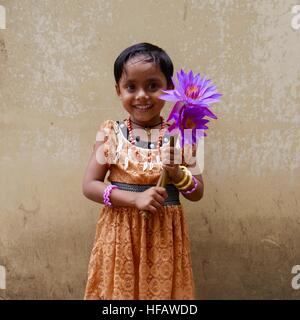 Sri Lankas Mädchen trägt einen ziemlich orange Kleid und Schmuck hält ein Angebot von zwei lila Lotusblumen Stockfoto