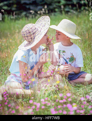 Glückliche Kinder, ein Junge und ein Mädchen, im Stroh Hüte sitzen in einem Feld riechen Wildblumen. Stockfoto