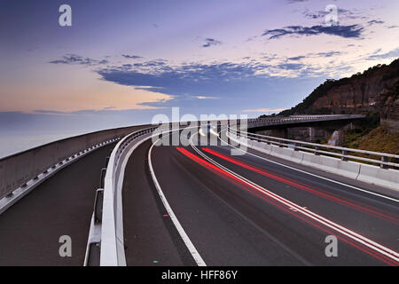 Australische Sea Cliff Bridge bei Sonnenuntergang am Great Pacific Drive malerischen touristischen Route in New South Wales. Verschwommen vorbeifahrenden Autos auf einer leeren Autobahn-Straße. Stockfoto