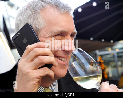 Lächeln entspannt Reifen City Geschäftsmann sitzt am Tisch im Restaurant im Freien, ein Gespräch auf seinem Smartphone iPhone 7 plus und trinken ein Glas Weißwein Stockfoto