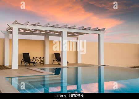 Weiße Pool-/ Pergola, dreht Pavillon bieten Schatten auf einer Terrasse Terrasse neben ein Infinity-Pool in der Dämmerung wie der Sonnenuntergang den Himmel Pinky ora Stockfoto