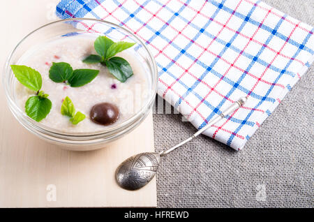 Nahaufnahme der Blick auf ein gesundes Gericht Haferflocken in eine Schüssel mit Glas Praline mit einem silbernen Löffel auf ein Holztablett und eine graue Tischdecke Stockfoto