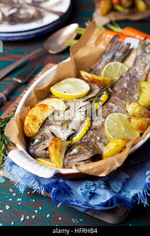 Ein Ofen gebratene Forellen in einem hohen Topf auf einem rustikalen Tisch. Mit Karotten und Kartoffeln auf der Seite. Stockfoto