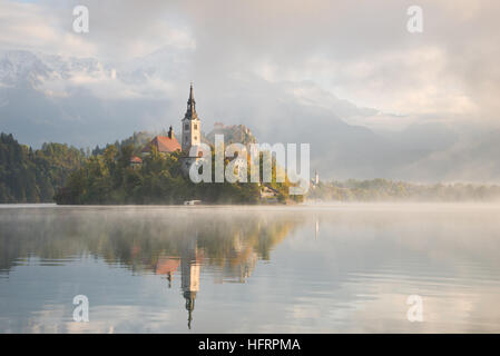 Kirche auf der kleinen Insel im Bleder See bei Sonnenaufgang an einem nebligen Morgen im Herbst mit Spiegelbild im Wasser in Slowenien. Stockfoto