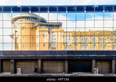 Ein Gebäudes spiegelt sich in den verspiegelten Fenstern eines anderen, als ein älterer Mann vorbei geht Stockfoto