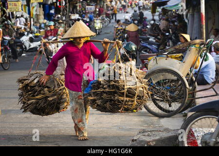 Barfuß Vietnamesisch Reife Frau in konischen asiatischen Hut tragen Holz an viel befahrenen Straße in der Nähe von chinesischen Viertels in My Tho, Vietnam. Stockfoto