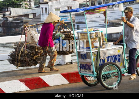 Barfuß Vietnamesisch Reife Frau in konischen asiatischen Hut tragen Holz an viel befahrenen Straße in der Nähe von chinesischen Viertels in My Tho, Vietnam. Stockfoto