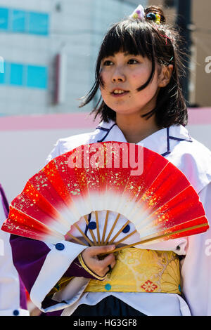 Japan, Kumamoto. Hinokuni Yosakoi Festival. Teenager-Mädchen Tänzer stehen mit leuchtend roten Fan eröffnet in der Taille. Im Freien, Sonnenlicht Stockfoto