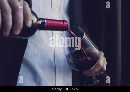 Horizontale Nahaufnahme von kaukasischen Mann im schwarzen Anzug und weißem Hemd strömenden Rotwein in ein hohes Glas in einer bar Stockfoto