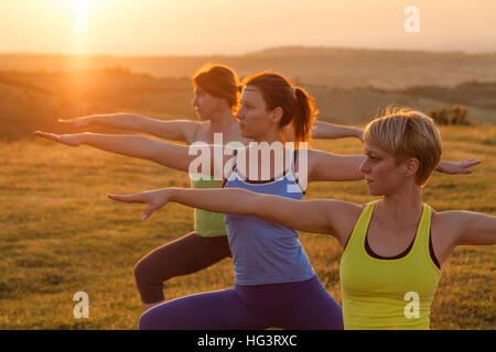 Drei Mädchen üben Yoga im Sonnenuntergang. Krieger-Position. Stockfoto