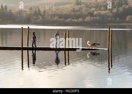 Zwei Personen mit einem Hund zu Fuß auf einem Holzsteg oder Liegeplatz auf Derwentwater Keswick Cumbria Uk im Wintersonne Stockfoto