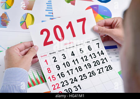 ein junger kaukasischer Geschäftsmann an seinem Schreibtisch voller Diagramme beobachtet einen Kalender 2017 Stockfoto
