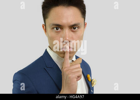 Menschliches Gesichtsausdruck. Junger asiatische Mann im blauen Anzug setzt Zeigefinger auf seine Lippen mit dem Ausdruck shh, ruhig, machen keinen Lärm.  Große Augen Kugel Mann. Weiß Stockfoto
