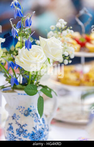 Weiße Rosen mit blauen Blumen und Babys Atem in einem blau-weißen Muster Porzellan Krug mit abgestuften Kuchenplatte Stockfoto