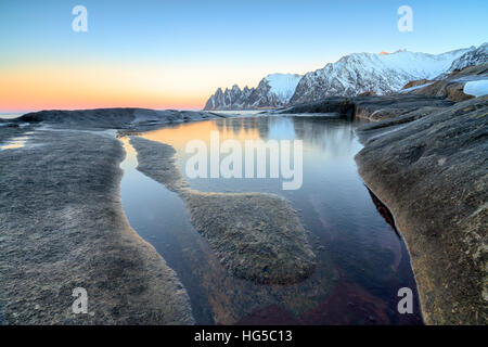 Orangefarbenen Himmel bei Sonnenuntergang spiegelt sich auf schneebedeckten Gipfeln und das gefrorene Meer, umgeben von Felsen Tungeneset, Senja, Troms Grafschaft, Norwegen Stockfoto