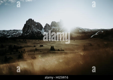 Holzhäuser auf einem Feld am Fuße einer Bergkette. Fotografiert bei Sonnenaufgang mit der Sonne als Hintergrundbeleuchtung. Stockfoto