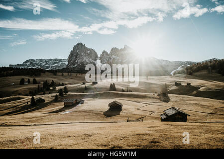 Holzhäuser auf einem Feld am Fuße einer Bergkette. Fotografiert bei Sonnenaufgang mit der Sonne als Hintergrundbeleuchtung. Stockfoto