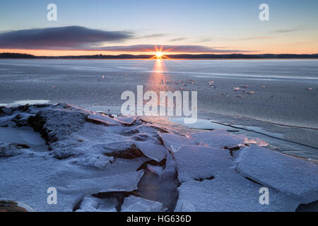 Sunrise und Gestein unter gestoßenem Eis auf einem gefrorenen See in Finnland im Winter. Stockfoto