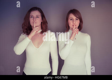 Doppelgänger / Alter Ego unter dem Motto Fotoshooting: zwei junge Frauen Mädchen tragen passende Kleidung Stand zusammenstehen passende Echo Spiegelung jedes andere körperliche Haltung und Ausdruck - ein Finger über den Mund - nichts Böses sprechen Stockfoto