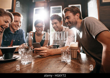 Gruppe von Freunden an einem Cafétisch sitzen und mit Blick auf Handy. Junge Männer und Frauen betrachten von Bildern auf Smartphone. Stockfoto