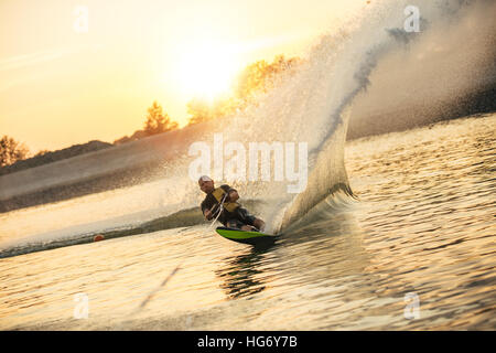 Wasserschifahrer bewegt sich schnell in Spritzwasser bei Sonnenuntergang. Mann Wakeboarden auf einem See Stockfoto