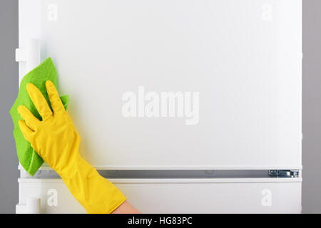 Zarte Frauenhand in schützenden gelben Latex Handschuh Reinigung weißer Kühlschrank mit grünes Tuch auf grauem Hintergrund Stockfoto