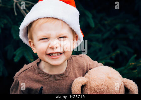 Porträt von fröhlicher Junge mit Teddy Bär trägt Weihnachtsmütze im Hinterhof Stockfoto