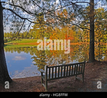 Alte hölzerne Parkbank sitzen an einem Teich im Herbst mit Bäumen mit drehen Blätter im Wasser gespiegelt. Stockfoto