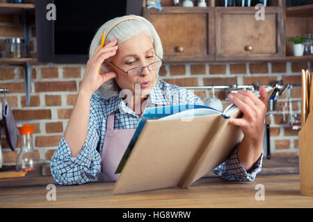 Ältere Frau in Brille lesen Kochbuch in Küche Stockfoto