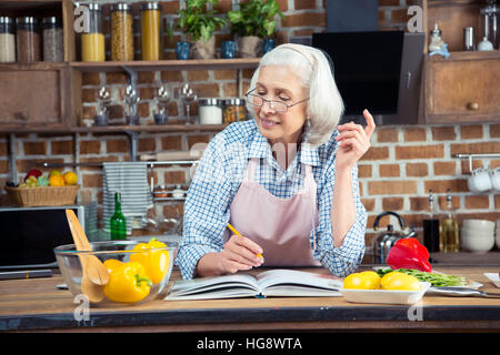 Lächelnde senior Frau schreiben in Kochbuch mit Bleistift und mit Blick auf die Inhaltsstoffe Stockfoto