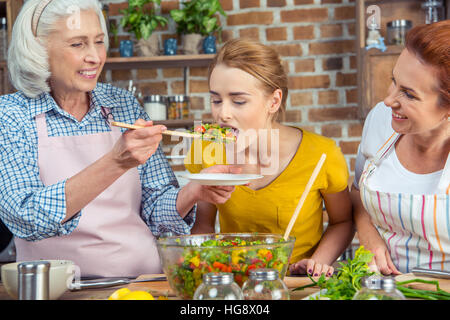 Junge Frau, die Verkostung von frischem Gemüsesalat mit Mutter und Großmutter in der Küche stehend Stockfoto