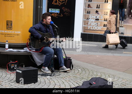 Junger Mann mit einer Gitarre, sitzend auf einer Bank im Fargate Sheffield Straßenmusik. Eine Frau kommt vorbei mit shopping im Hintergrund Stockfoto