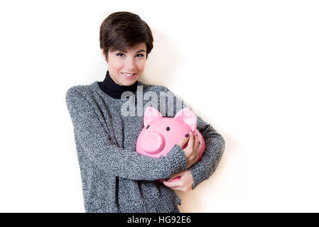 Junge Frau glücklich umarmt ein Sparschwein mit Einsparungen, Blick in die Kamera und lächelnd auf weißem Hintergrund isoliert textfreiraum auf der rechten Seite Stockfoto