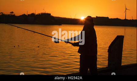 Ein Fischer bei Sonnenuntergang wirft seine Rute und Schnur in der Hoffnung auf einen Fisch fangen. Die Sonne ist fast verschwunden, da das letzte Licht ist Stockfoto