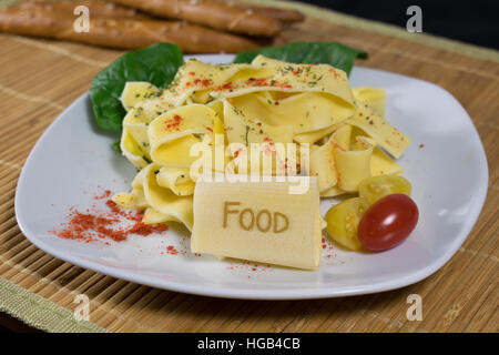Nudelgericht gekleidet auf einem weißen Teller mit Blattspinat und Tomaten dekoriert. Auf der vorderen Noodle ist das Wort Essen. Stockfoto