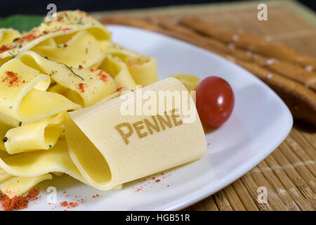 Nudelgericht gekleidet auf einem weißen Teller mit Blattspinat und Tomaten dekoriert. Auf der vorderen Noodle ist das Wort Penne. Stockfoto